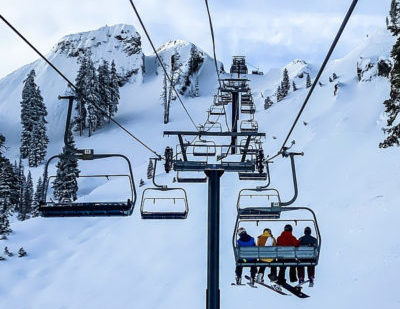 Four-person ski lift in winter looking up a snow-covered mountain. One chair is filled with a combination of four skiers and snowboarders riding up the mountain, with their backs facing the camera.